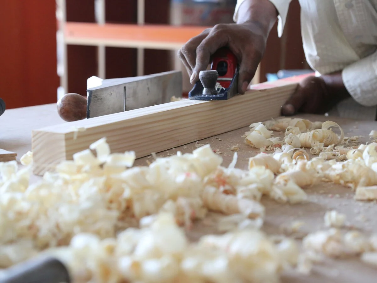 A worker guiding a hand plane along a long pale timber plank on a workbench, a pile of wood shavings in the foreground, the workshop behind out of focus.