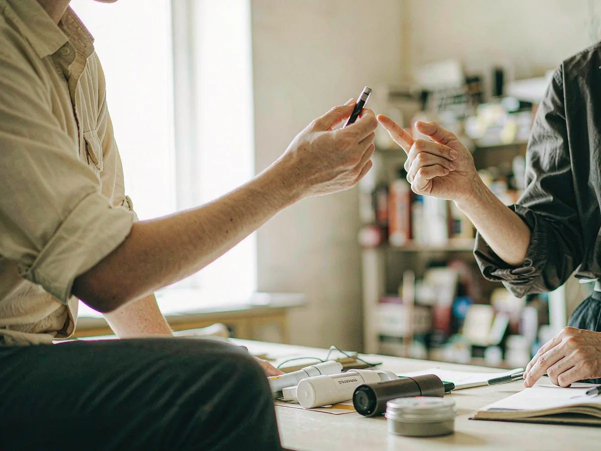 A buyer and a supplier representative comparing physical product samples across a work table, one sample held up to the light.