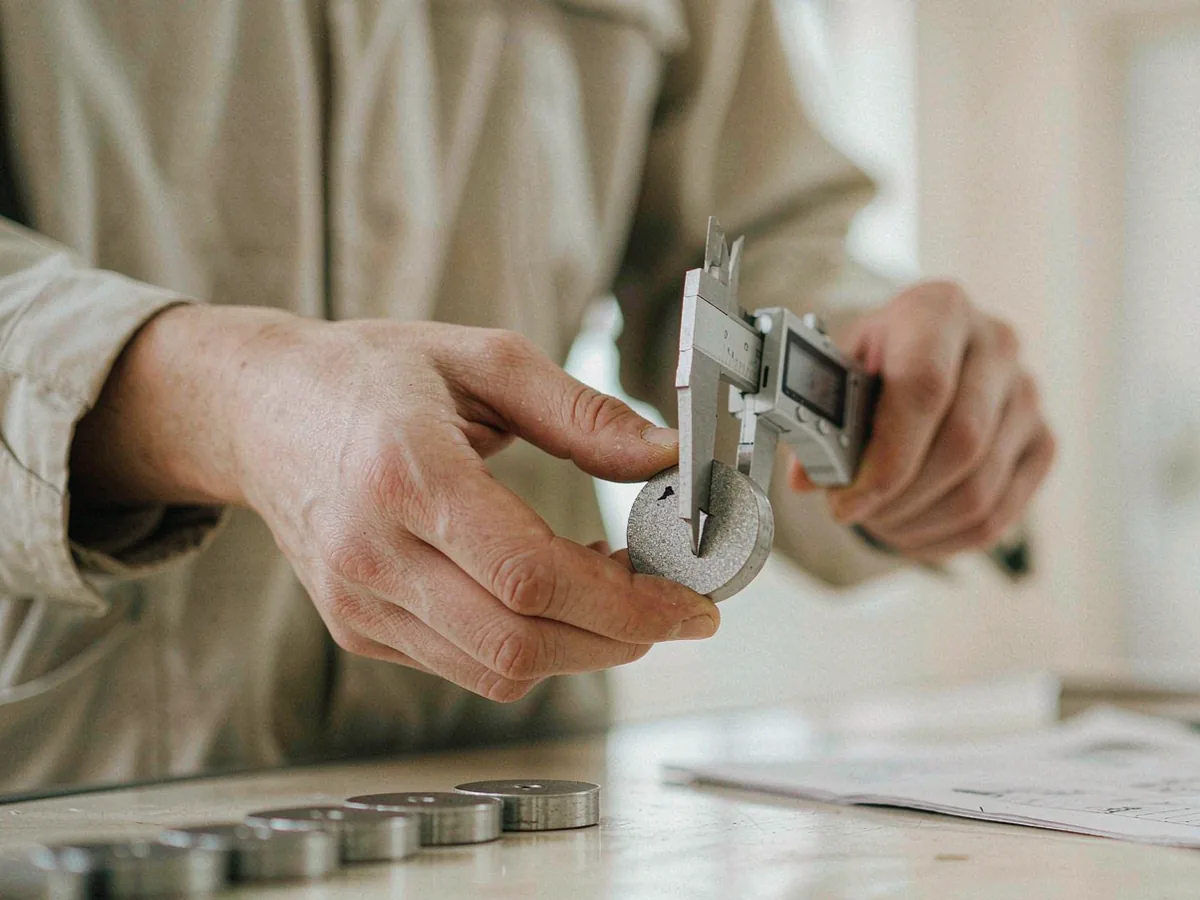 Close view of an inspector measuring a small metal component with a digital caliper, identical parts lined up beside.
