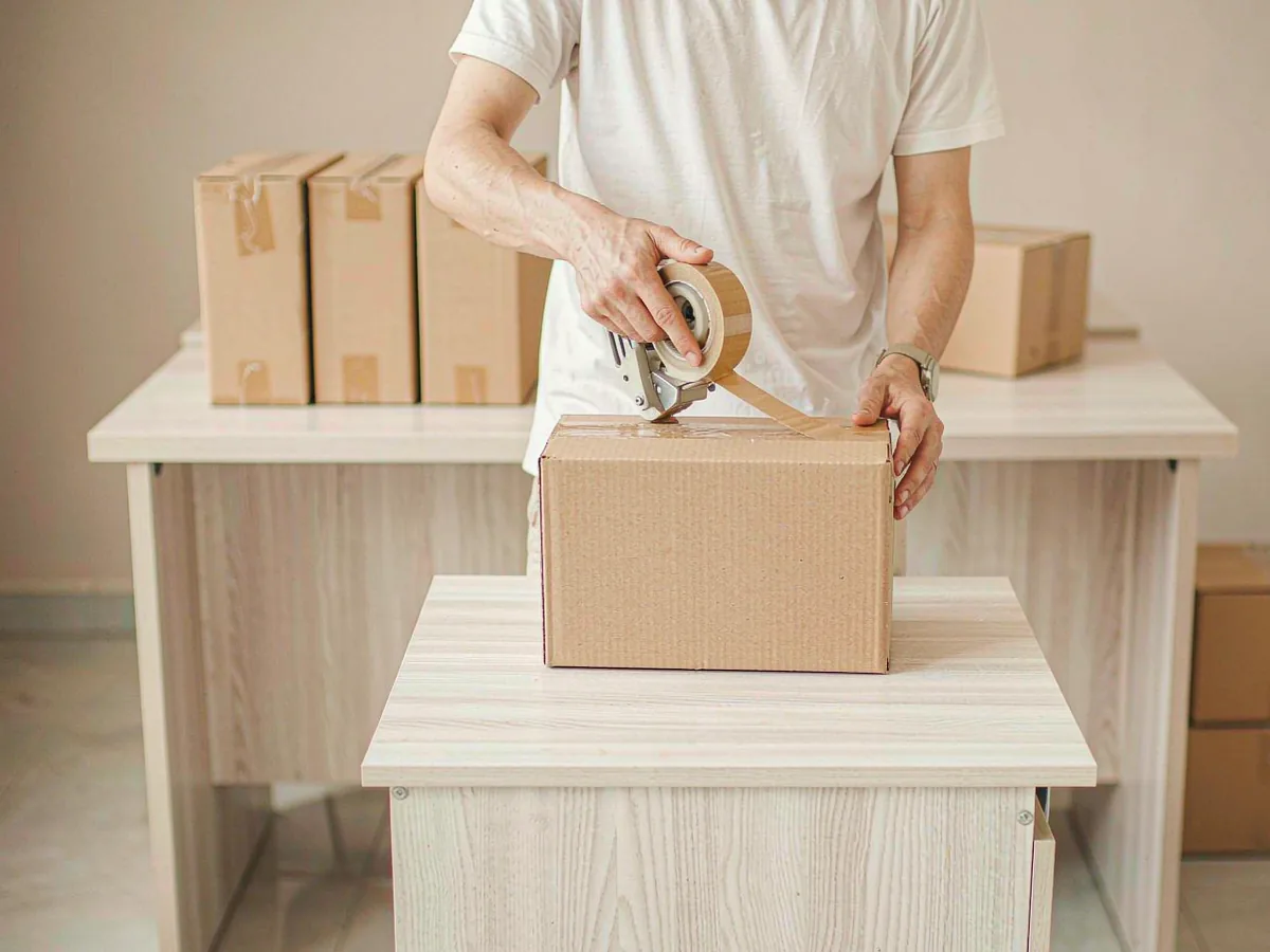 A worker sealing a plain cardboard carton with a tape dispenser on a packing table, more cartons blurred behind.