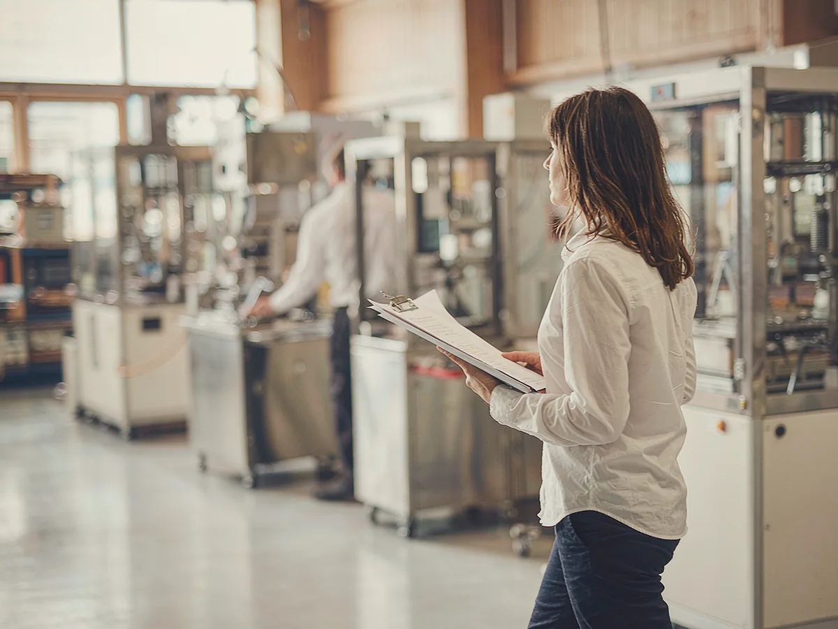An assessor walking through a factory floor with a clipboard, machinery softly out of focus behind her.