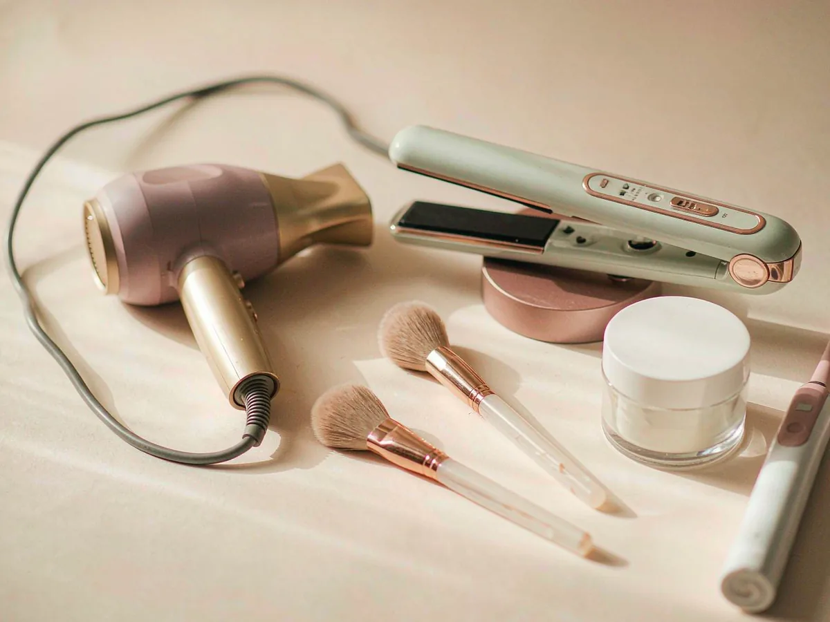 A hair dryer, a flat iron, makeup brushes and a jar of cream arranged together on a pale surface.
