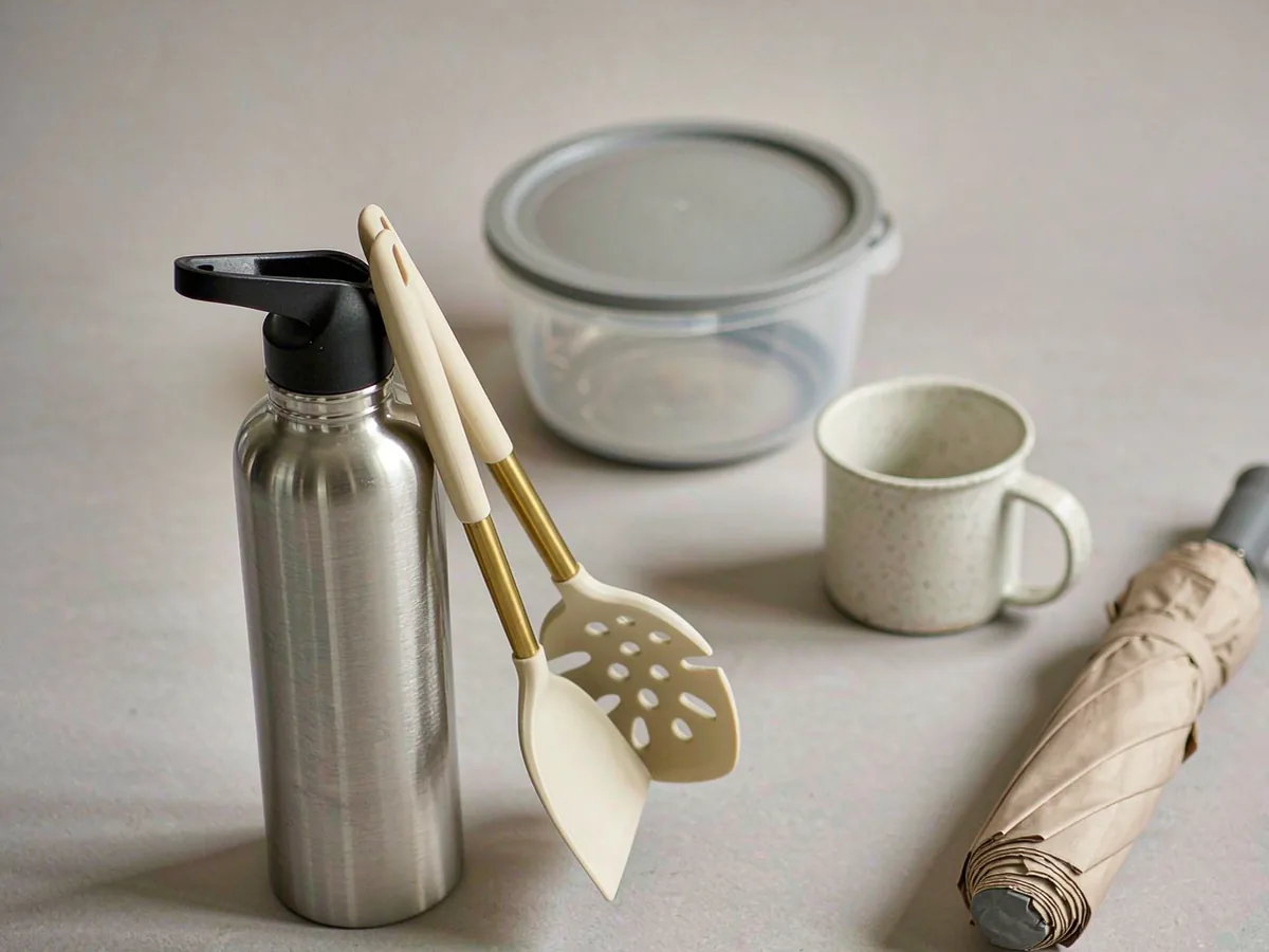 A stainless insulated bottle, silicone kitchen utensils, a lidded container, a ceramic mug and a folded umbrella arranged together on a pale surface.