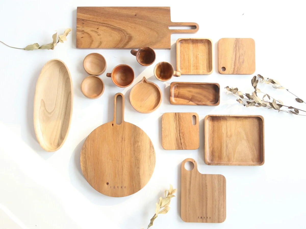 A flat lay of wooden kitchenware on a pale background — cutting boards, serving trays, small bowls and cups in assorted shapes and sizes, with dried stems at the edges.